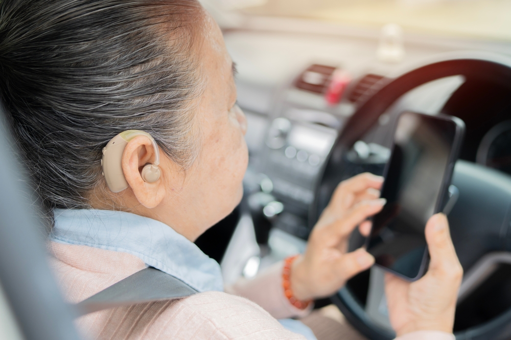 An older woman with a hearing aid is sitting in the driver’s seat of a car, wearing a seatbelt, and looking at a smartphone she is holding in her right hand. The interior of the car is visible.