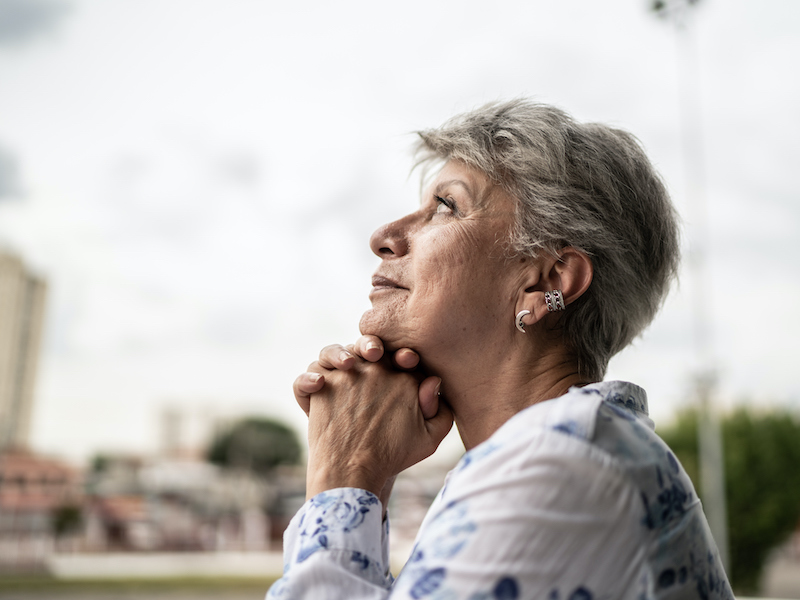 An older woman with short gray hair gazes upward thoughtfully, resting her chin on her clasped hands. She wears a light, patterned blouse and is outdoors with blurred buildings and greenery in the background.
