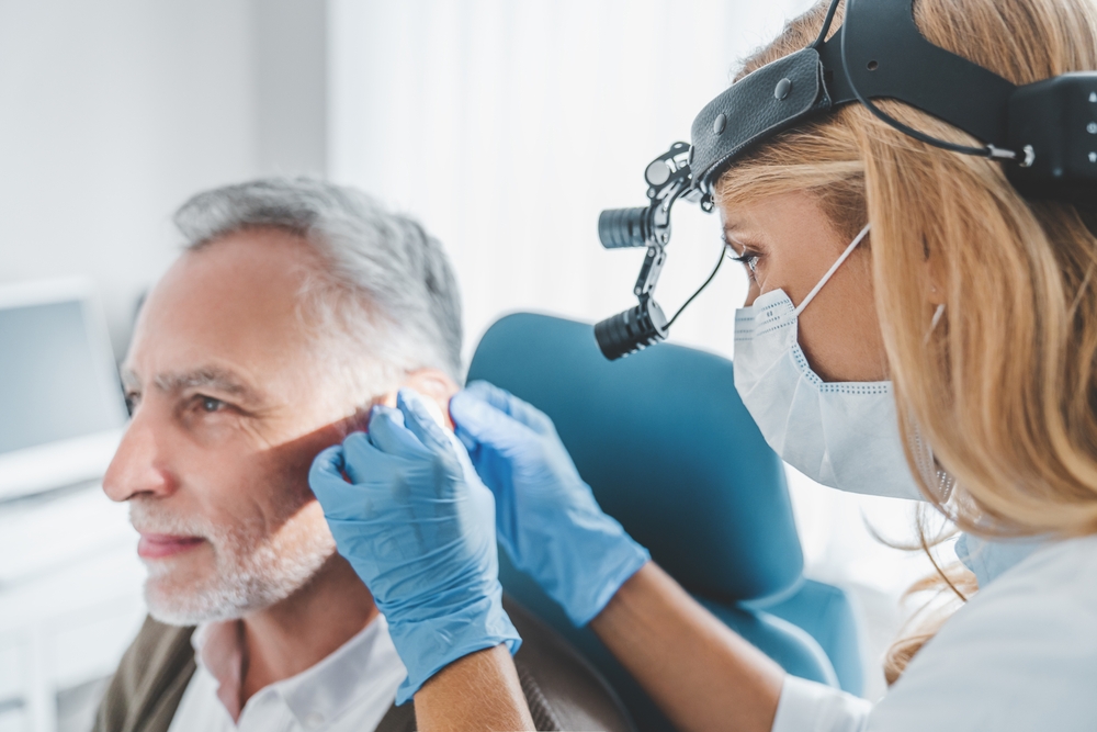 A female doctor wearing a mask and headlamp examines an older mans ear with a medical instrument in a clinic. The man sits calmly while the doctor works carefully, both in a well-lit medical office.
