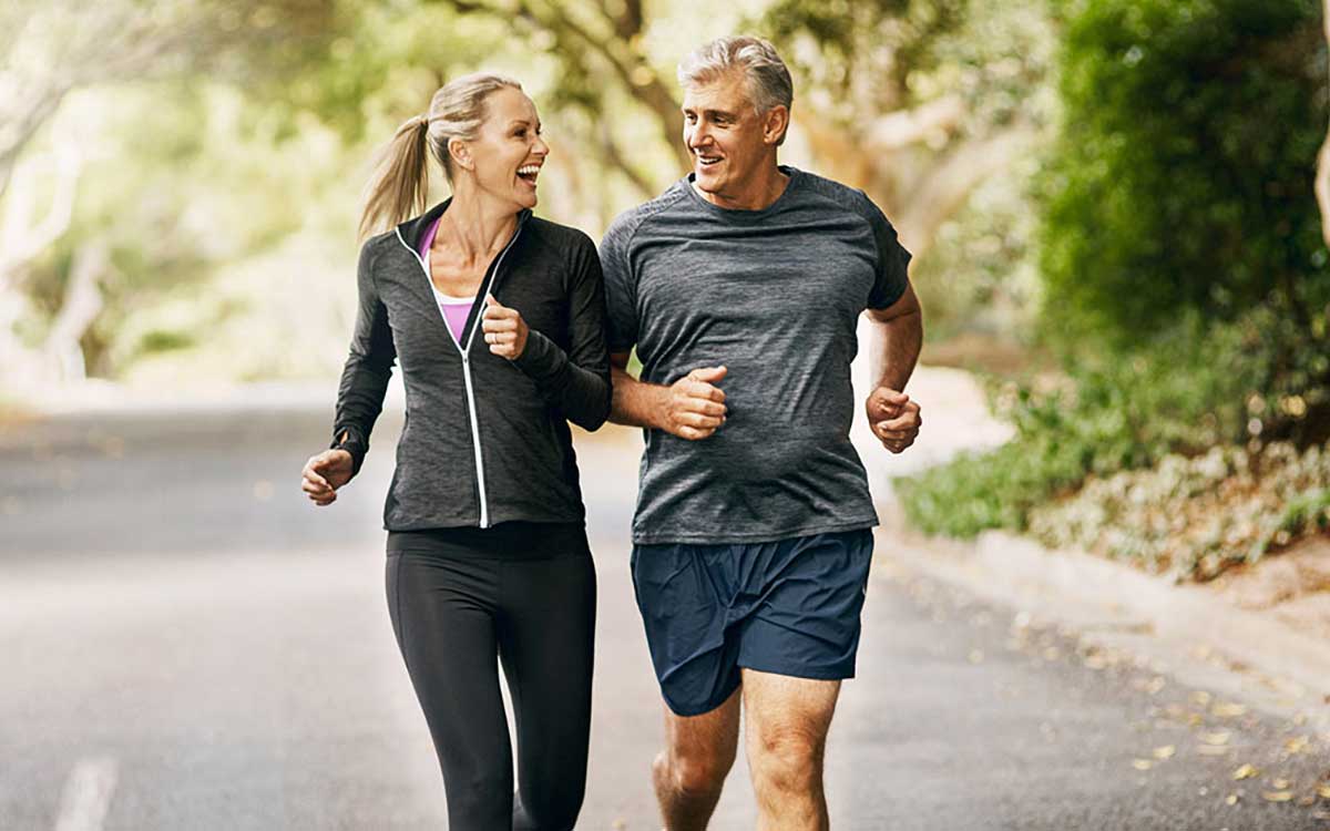 A smiling middle-aged couple jogs together outdoors on a tree-lined path, wearing athletic clothing and enjoying each others company on a sunny day.