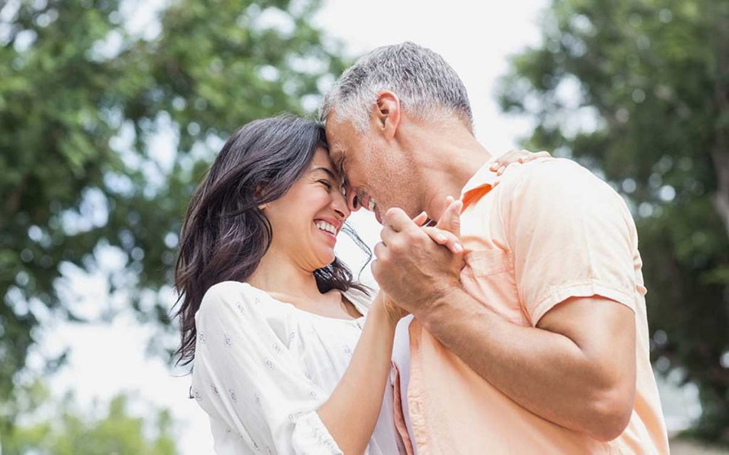 A smiling couple embraces outdoors, holding hands and touching foreheads, with trees blurred in the background. They appear happy and affectionate, enjoying a moment together.