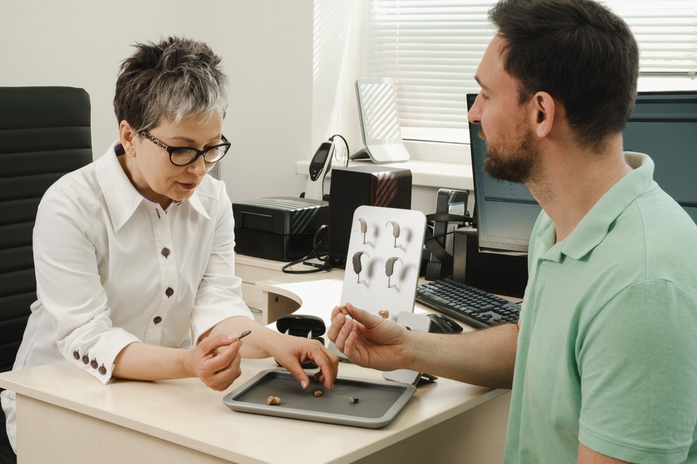 A woman in glasses shows different hearing aids to a seated man in a green shirt at a desk in a modern office, possibly during a hearing aid consultation.