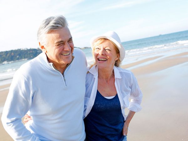 An older couple walks on the beach, smiling and looking at each other. The man wears a white sweater, and the woman wears a white shirt and hat. The ocean and blue sky are in the background.