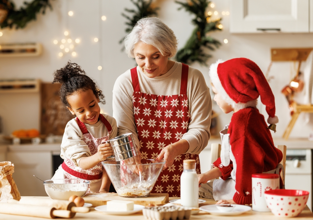 A woman and two children, all wearing festive aprons, bake together in a decorated kitchen. One child wears a Santa hat and the group smiles while mixing ingredients in a bowl. Holiday decorations are visible in the background.