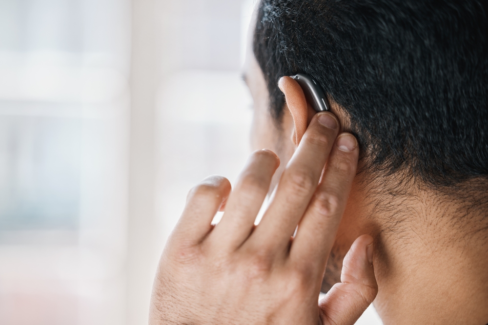 A person with short dark hair adjusts a hearing aid behind their right ear using their right hand, shown from the back in close-up.