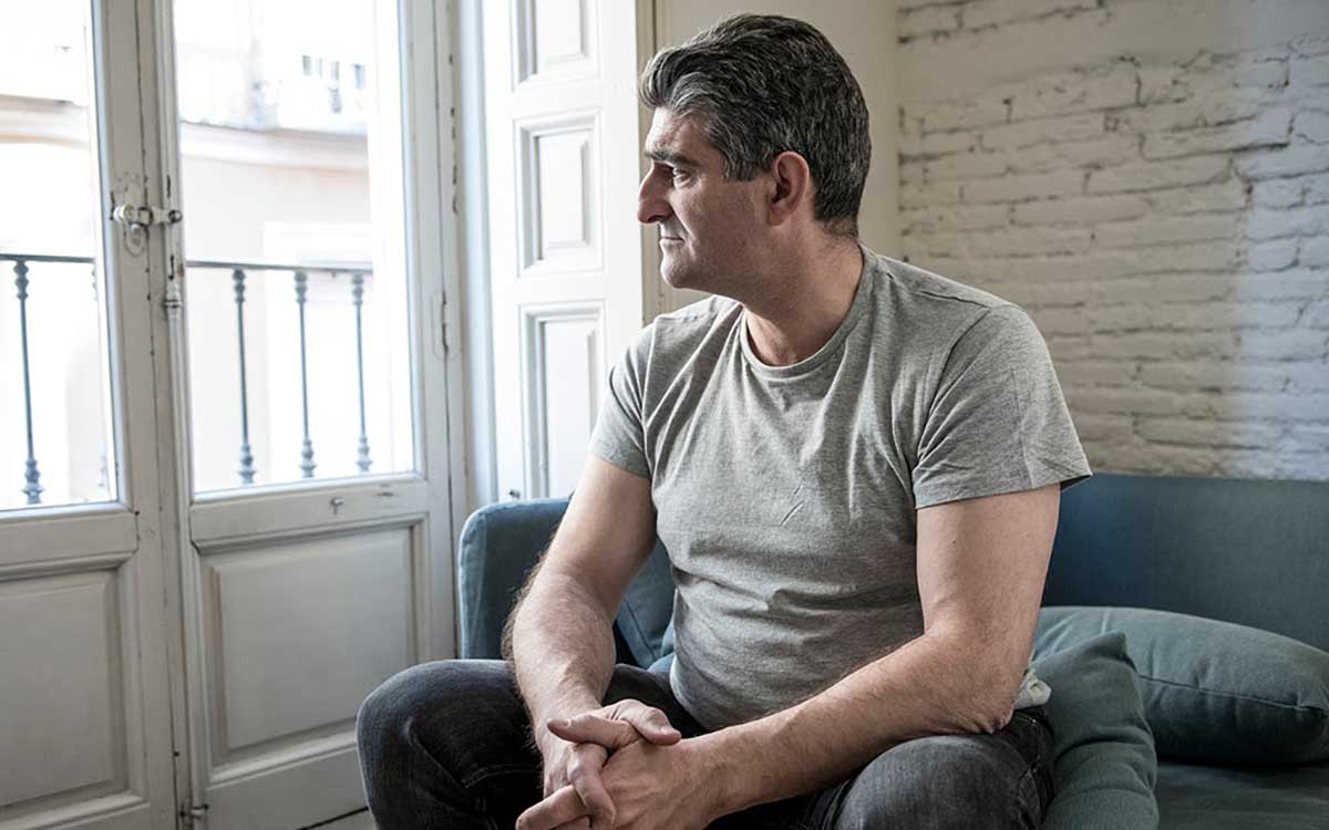 A man with gray hair in a gray t-shirt sits on a couch indoors, looking out a window with a thoughtful expression. Brick and white painted walls are visible in the background.