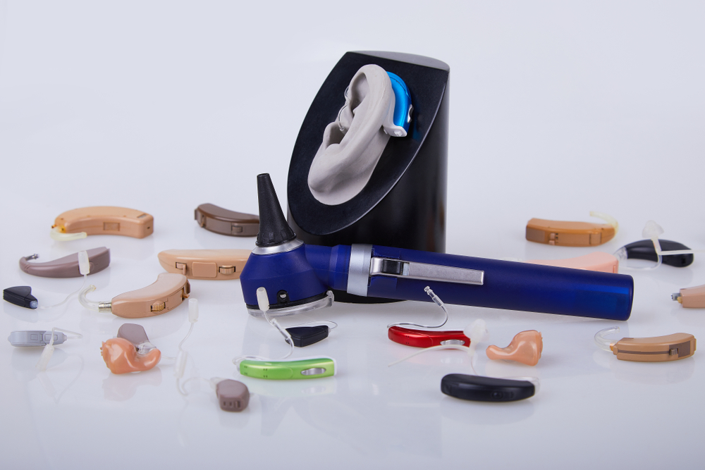 A variety of hearing aids in different shapes and colors surround an otoscope and an ear model with a blue hearing aid, all displayed on a white surface.