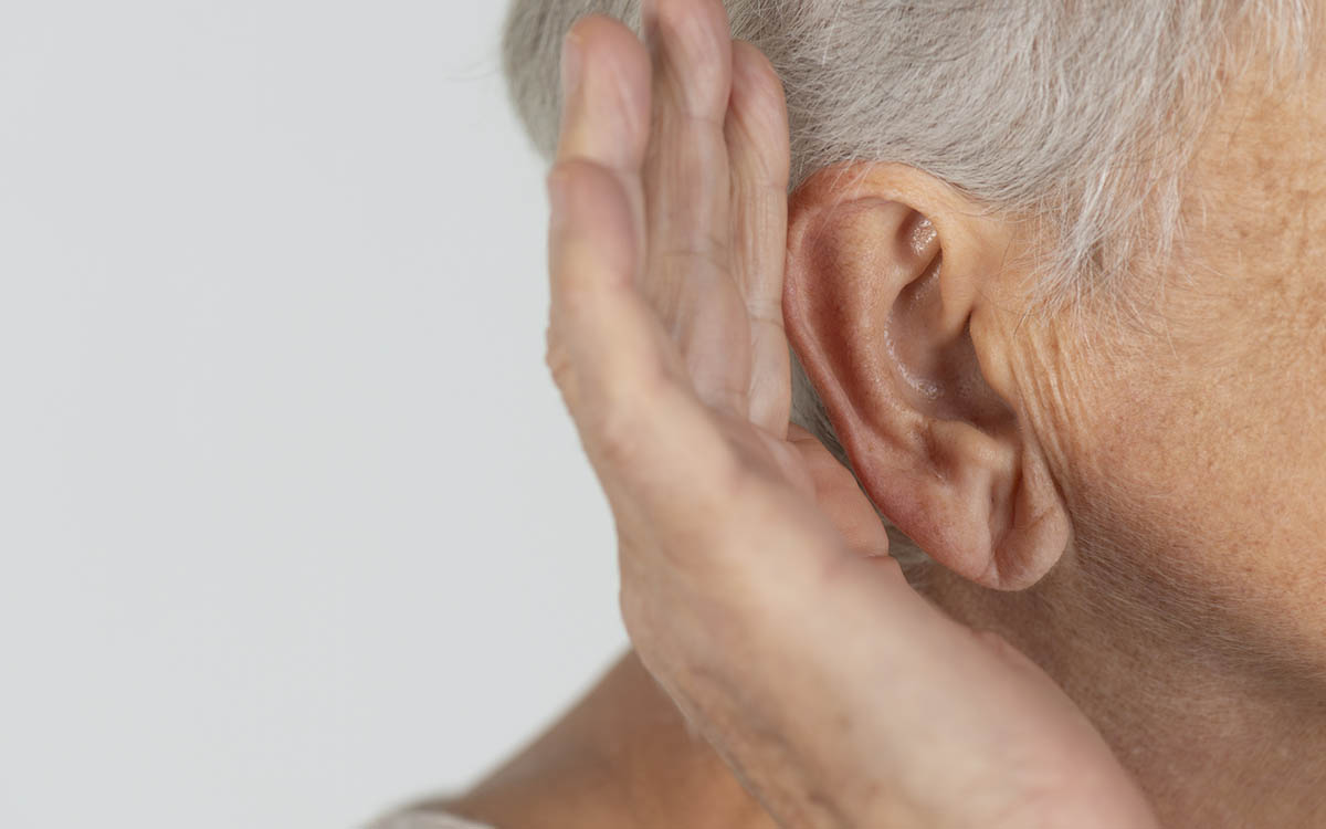Close-up of an older adult’s ear with their hand cupped behind it, as if straining to hear better, against a plain light background.