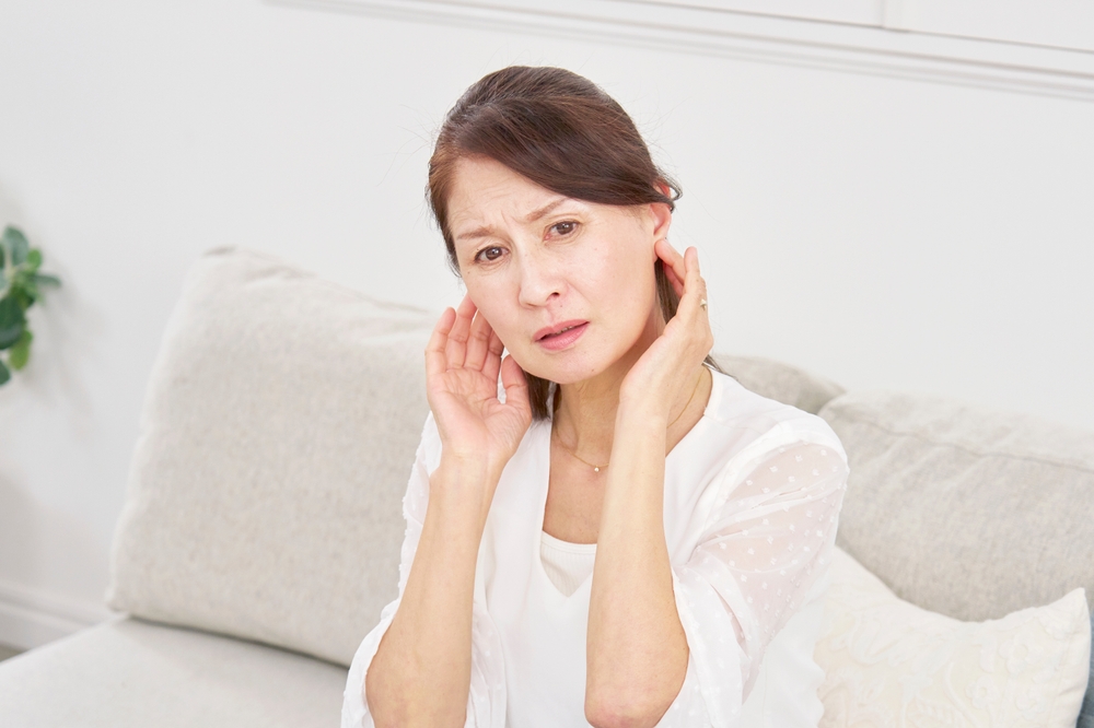 An older woman sitting on a couch touches both ears and looks concerned or uncomfortable, as if experiencing ear pain or discomfort.