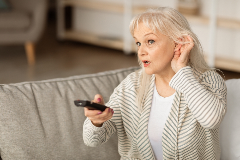An older woman sits on a couch holding a TV remote in one hand and cupping her ear with the other, appearing to have difficulty hearing the television.