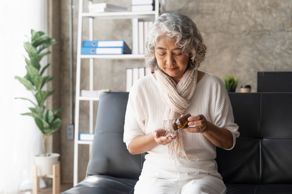 An older woman with gray hair sits on a black sofa, wearing a light scarf and white clothes, pouring pills from a brown bottle into her hand. A bookshelf and a potted plant are in the background.