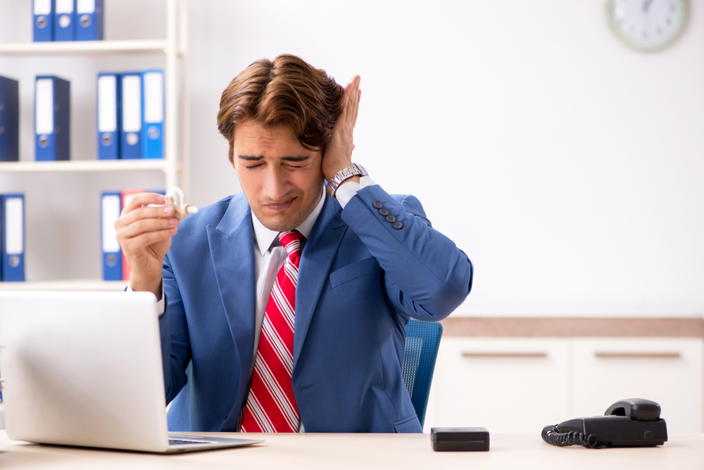 A man in a suit sits at a desk with a laptop, holding a cup and covering one ear, appearing uncomfortable or in pain. Office supplies and a phone are on the desk, and shelves are in the background.