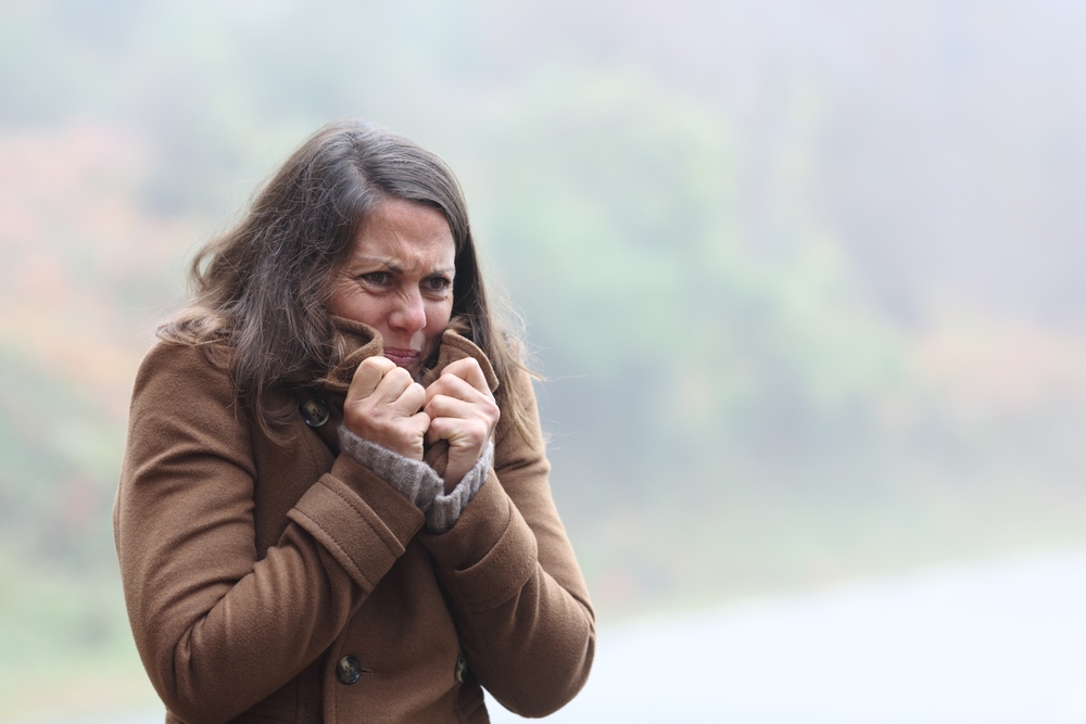 A woman wearing a brown coat stands outdoors, holding her collar up and shivering from the cold, with a blurred, misty background of trees and grass.