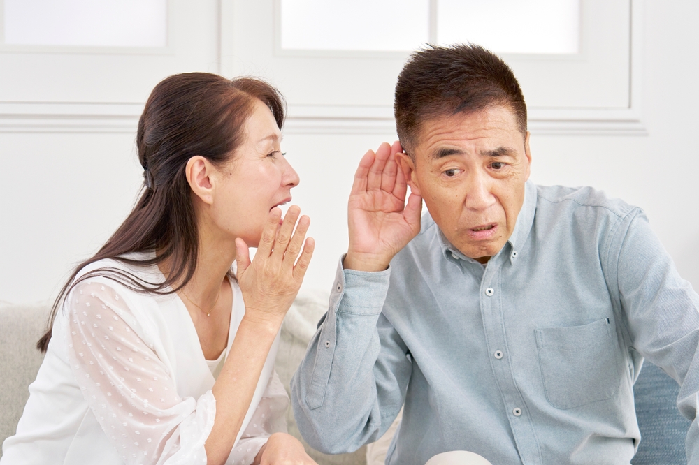 A woman is whispering to a man, who is leaning in with his hand to his ear, appearing to listen closely. Both are indoors, sitting on a couch, and seem to be having a private or secretive conversation.