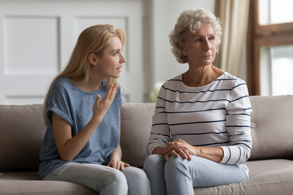 A younger woman appears to be speaking emotionally to an older woman who is looking away with a serious expression. They are sitting together on a couch in a well-lit living room.