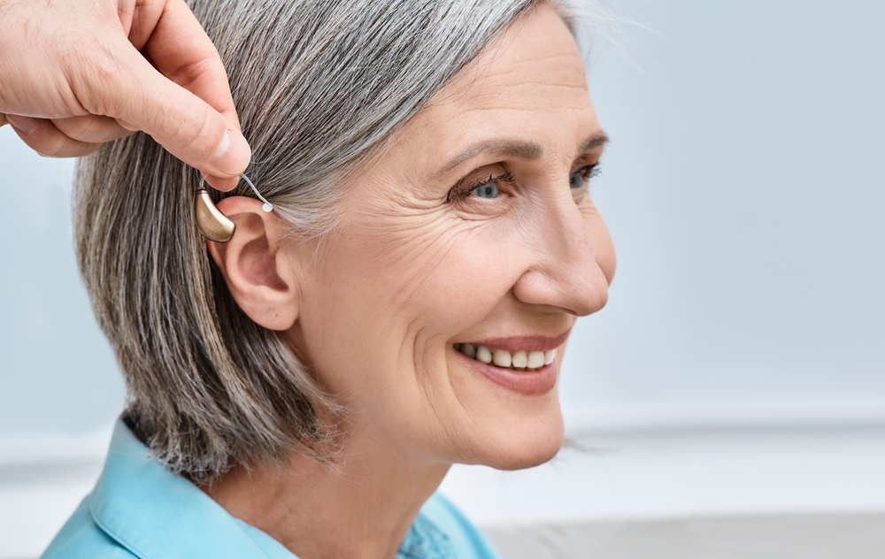 An older woman with gray hair smiles as someone fits a behind-the-ear hearing aid onto her ear. She is wearing a light blue shirt and appears happy and relaxed.