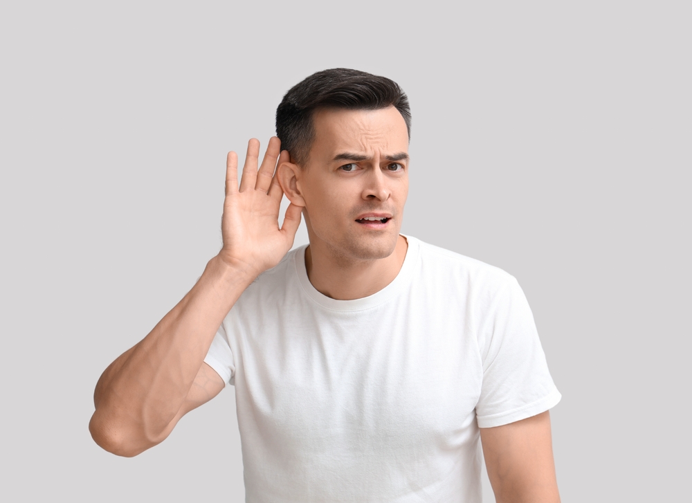 A man in a white t-shirt holds his hand to his ear and looks confused, as if trying to hear something better. The background is plain light gray.