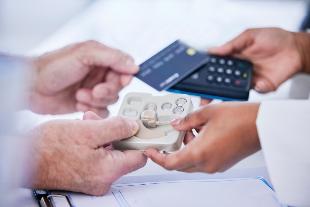 Close-up of two people exchanging a payment card and a point-of-sale terminal, while holding a small package containing a hearing aid device on a tray.