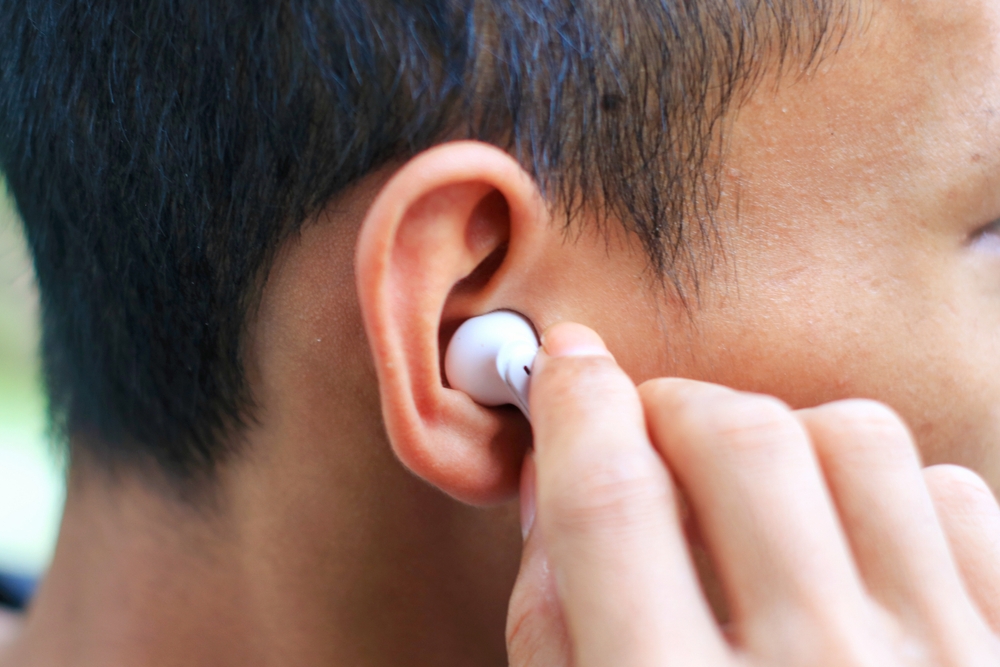 A close-up of a person’s ear with short black hair, inserting a white wireless earbud using their right hand.
