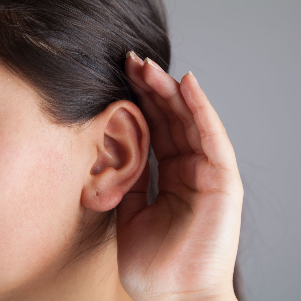 A person holds a hand behind their ear, appearing to listen closely. The focus is on the ear and hand, with a neutral gray background.