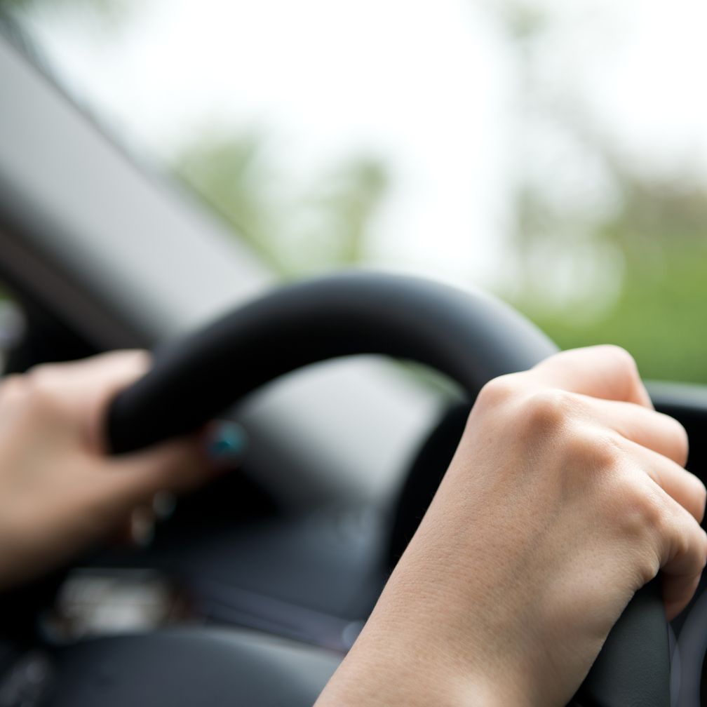 Close-up of a person’s hands gripping a steering wheel while driving a car, with a blurred background visible through the windshield.