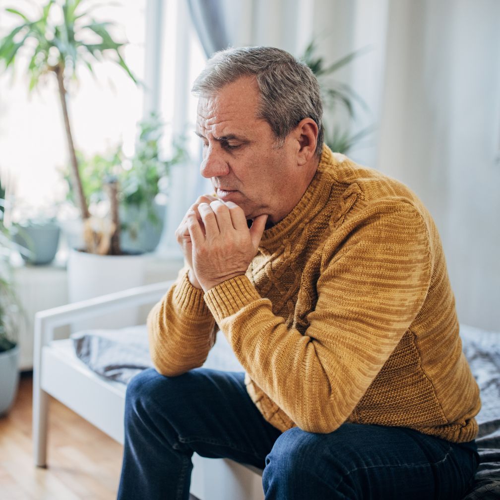 An older man in a mustard sweater sits on the edge of a bed, looking thoughtful and concerned. Houseplants and a window are visible in the background.