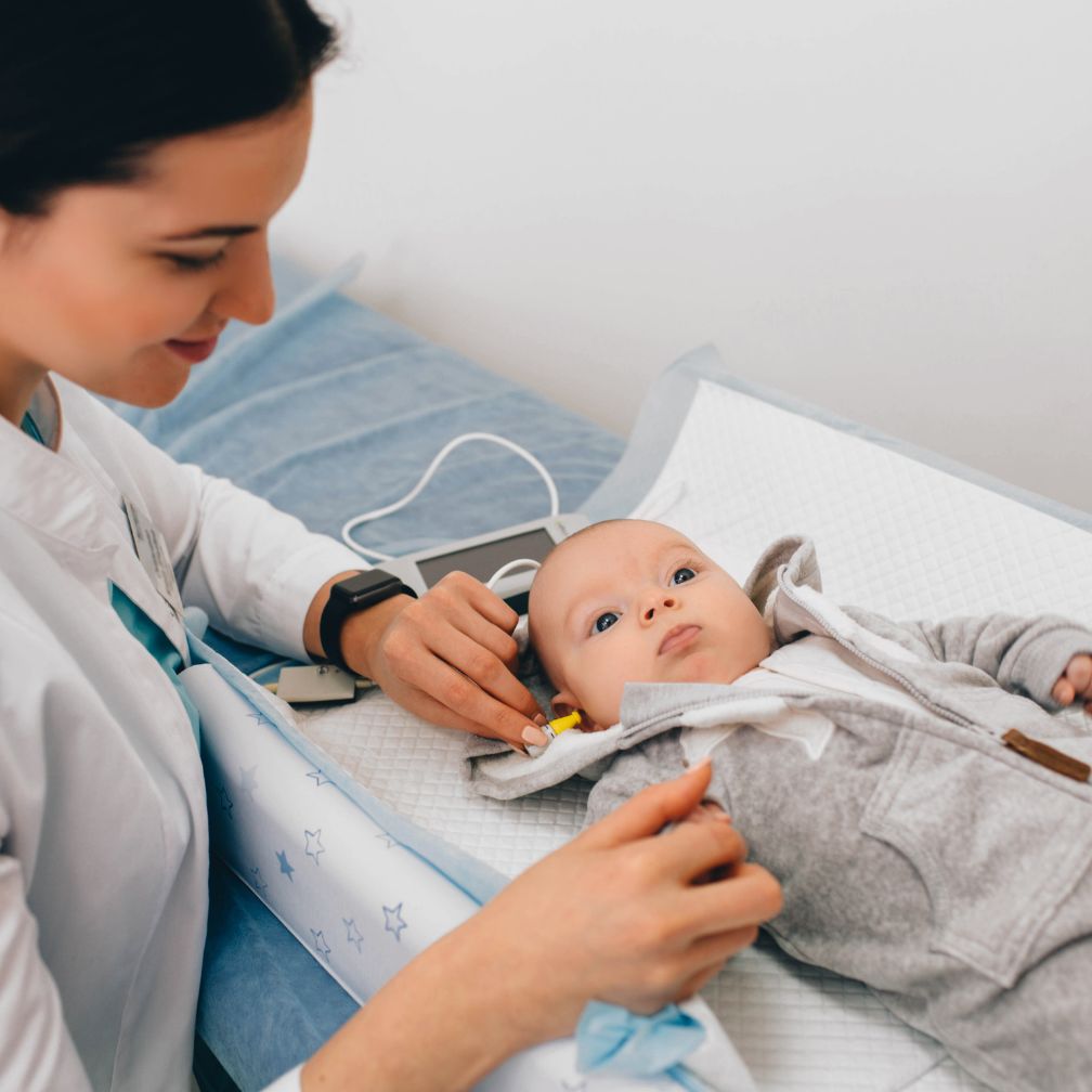 A healthcare professional tests a babys hearing using a small device in the babys ear. The baby lies on an exam table, looking up, while the professional smiles and gently holds the equipment.