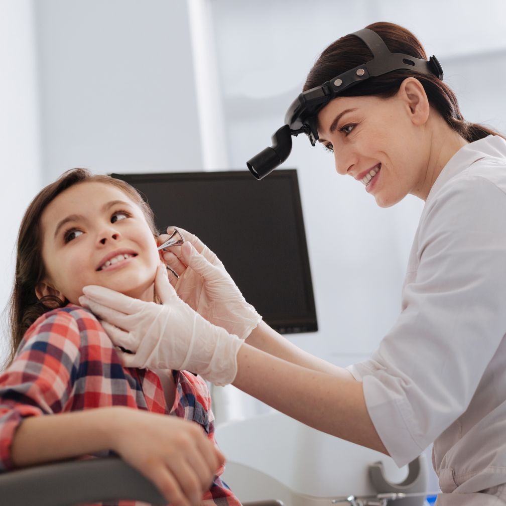 A doctor wearing gloves and a headlamp examines a young girls ear with an otoscope in a medical office. The girl is smiling and wearing a plaid shirt.