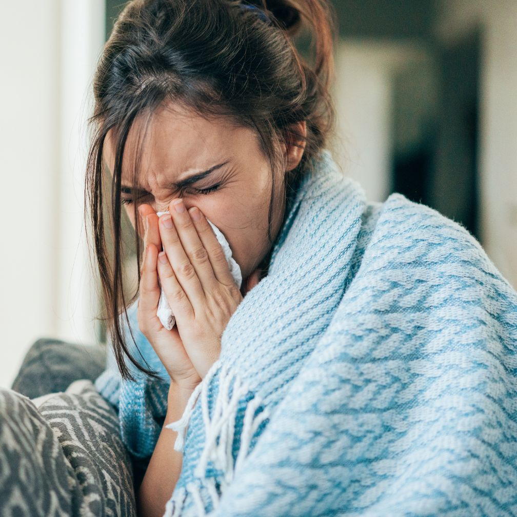 A person wrapped in a blue blanket blowing their nose into a tissue.