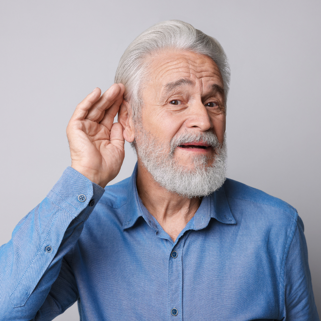 An older man with a white beard wearing a blue button-down shirt cups his hand behind his ear to listen.